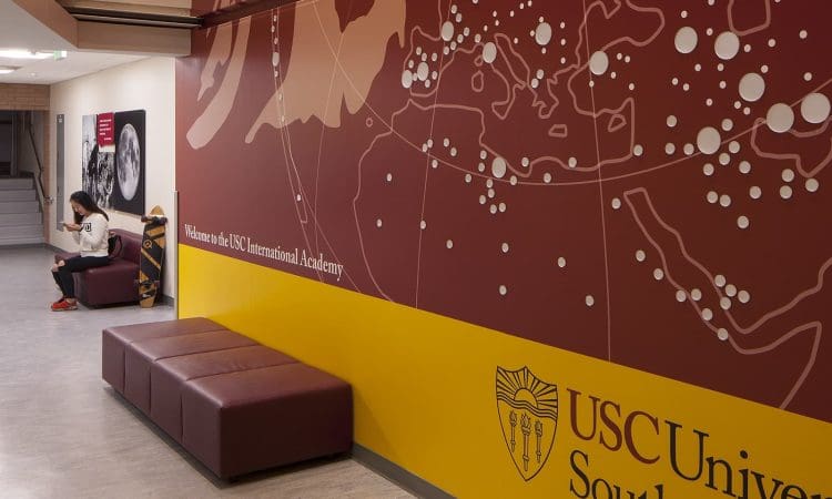 A student sits on a bench in a hallway with a large USC International Academy world map mural on the wall.