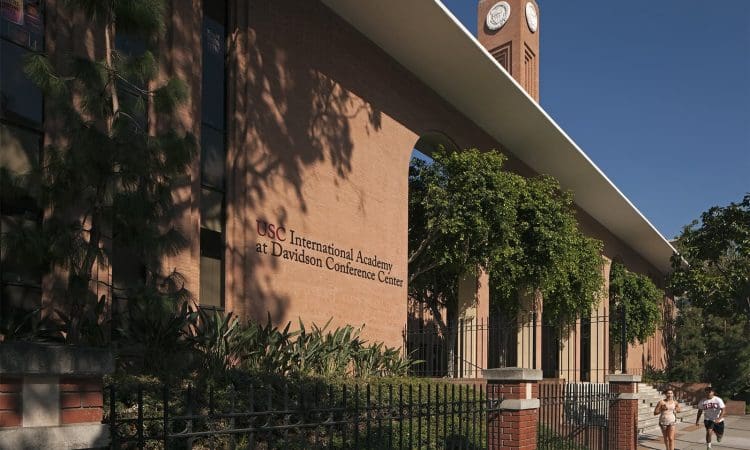 Red brick building with "USC International Academy at Davidson Conference Center" on the facade, clock tower in the background, trees and two people walking on the sidewalk.