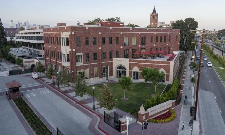 Aerial view of a multi-story red-brick building on a neatly landscaped campus with a clock tower in the background and surrounding roads visible.