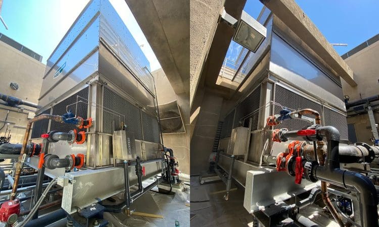 Two large industrial HVAC units with multiple pipes and valves are installed outdoors between concrete walls under a clear blue sky.