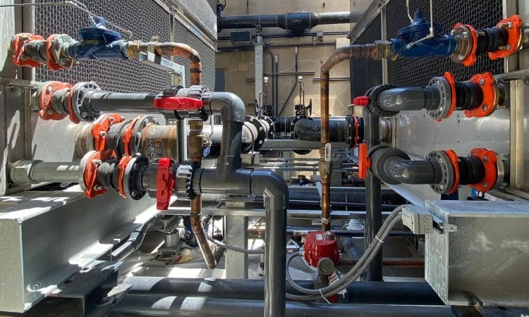 Industrial piping system with multiple valves, connectors, and metal pipes, featuring red and blue valve handles, set between two large units.