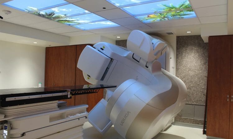 A modern medical imaging or radiotherapy machine is positioned in a treatment room with a ceiling display of blue sky and palm trees.