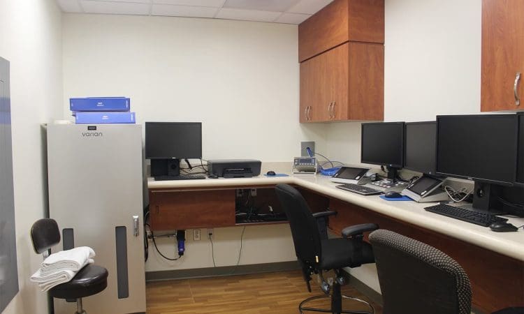 Small office with multiple computer monitors on a desk, office chairs, a large equipment cabinet, and wooden cabinets mounted on the wall.