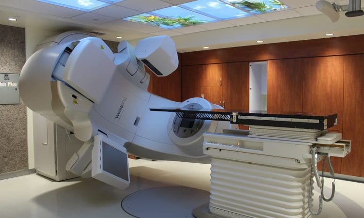 A medical linear accelerator machine in a modern treatment room with wood-paneled walls and a ceiling featuring a sky and tree design.