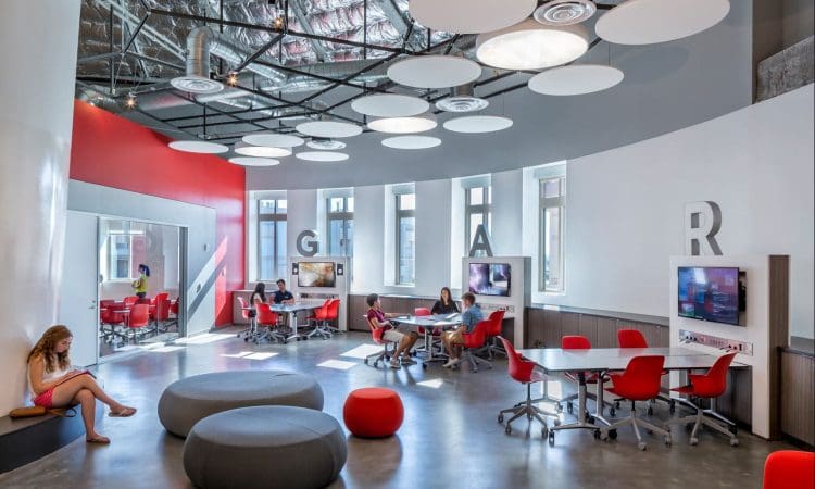 Modern classroom with red chairs, round gray seating, wall-mounted monitors, and students working in groups; exposed ceiling and large letters on walls.