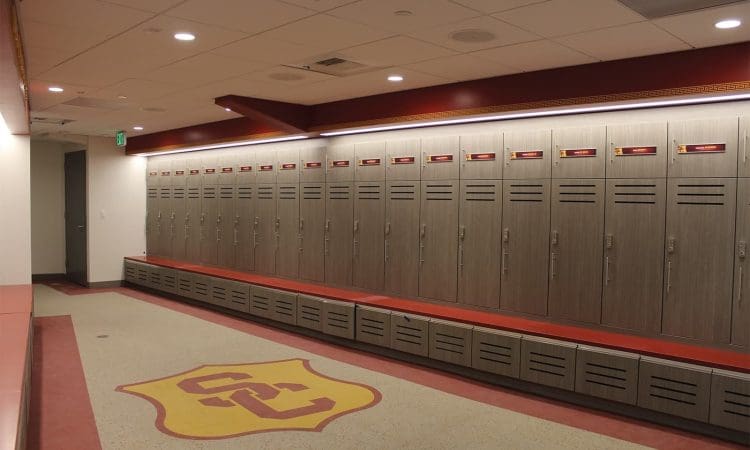 A row of wooden lockers lines the wall of a locker room with red and gold accents and a large USC logo on the floor.