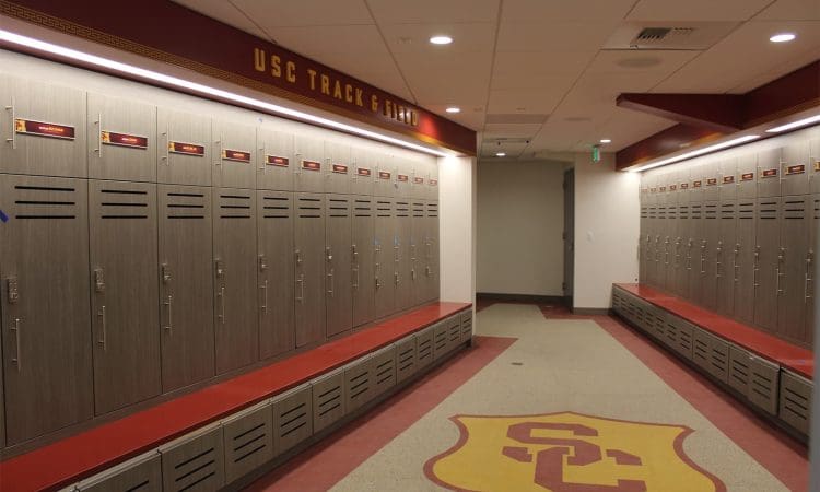 Empty locker room with gray lockers and red benches, "USC Track & Field" sign above, and large USC logo on the floor.