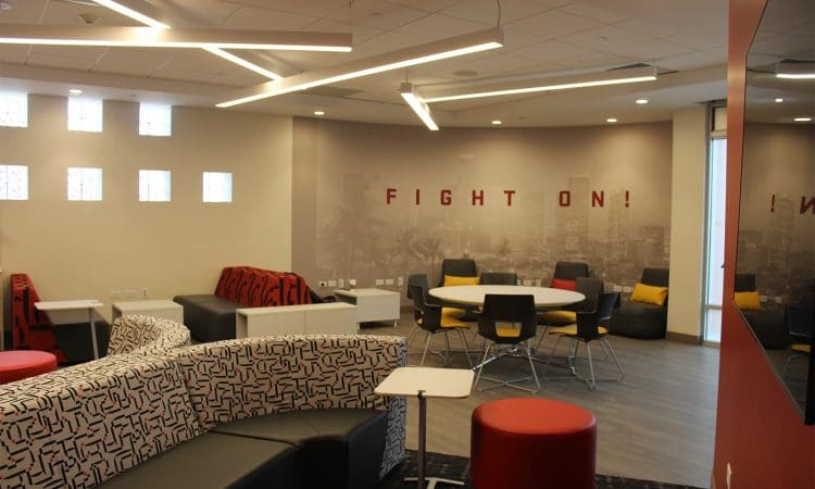 Modern lounge area with patterned seating, round tables, chairs, and “FIGHT ON!” on the wall, illuminated by geometric ceiling lights.