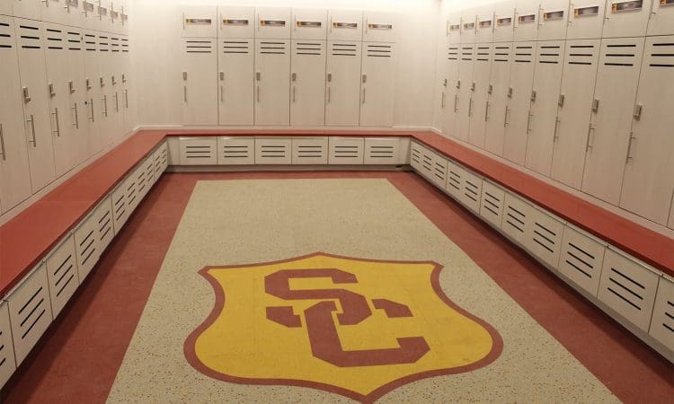 Empty locker room with white lockers, red benches, and a large yellow and red "SC" logo on the floor in the center of the room.