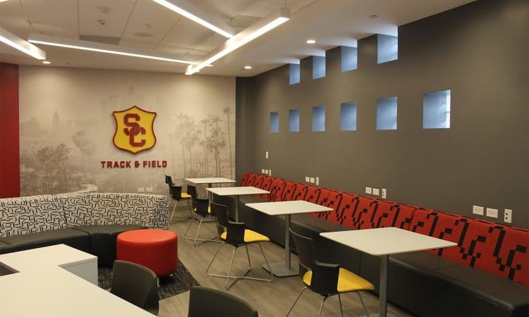 Modern lounge area with tables, chairs, and a long bench featuring USC Track & Field branding on the wall and square windows on a gray accent wall.
