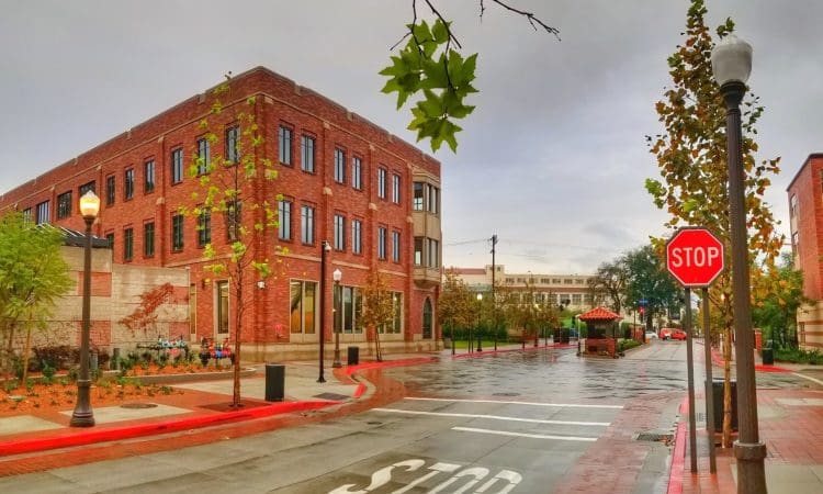 A quiet, empty street with red brick buildings, trees, a stop sign, and wet pavement on a cloudy day.