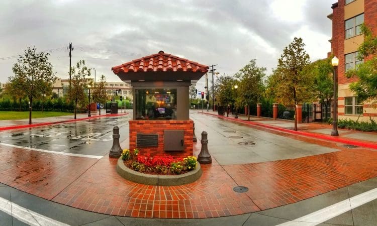 A small brick security booth with a red tile roof is centered at the entrance to a quiet, wet street lined with trees and buildings under a cloudy sky.