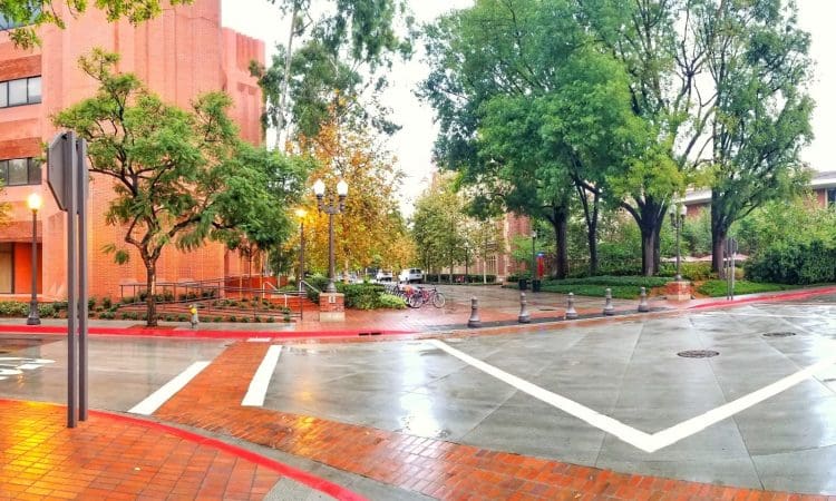 A quiet, wet intersection on a college campus with red brick buildings, trees, bike racks, and street lamps on a cloudy day.