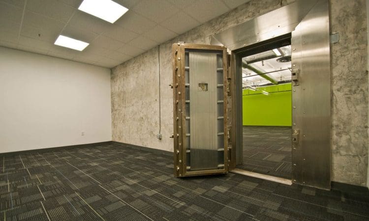 A large, open vault door leads from a carpeted room with plain walls to a room with bright green walls and exposed ceiling pipes.