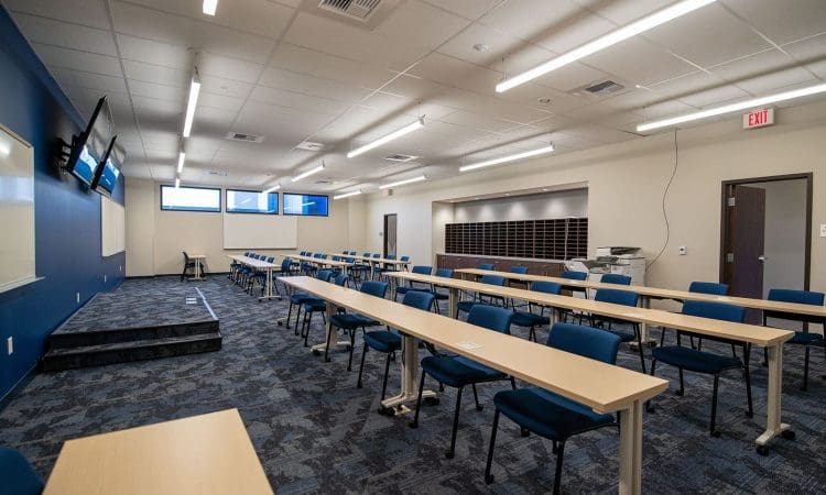 Modern classroom with blue chairs and long tables facing a wall-mounted screen and whiteboard; windows provide natural light, and a mail sorter is visible at the back.