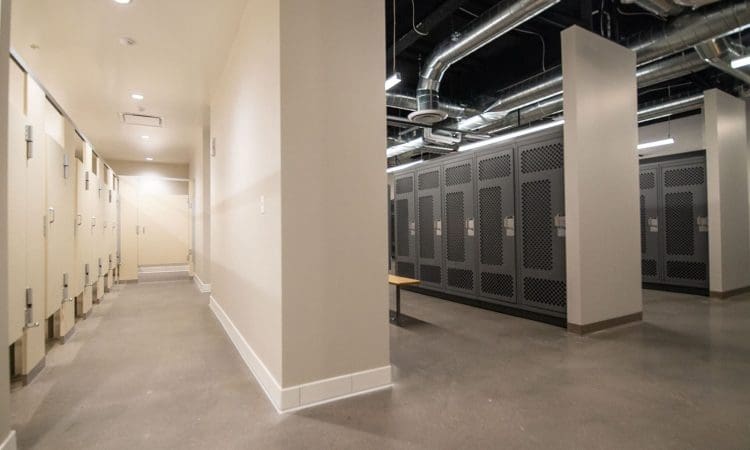 A clean, modern locker room with beige walls, gray locker units, overhead ducts, and a row of restroom stalls on the left.