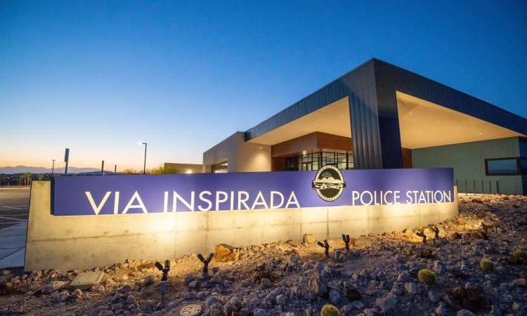 A modern building with a sign reading "Via Inspirada Police Station" illuminated at dusk, with desert landscaping in the foreground.
