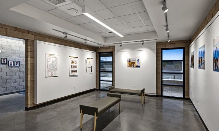 Modern art gallery with white walls, framed artworks, two benches in the center, track lighting, and large windows showing an outdoor area.