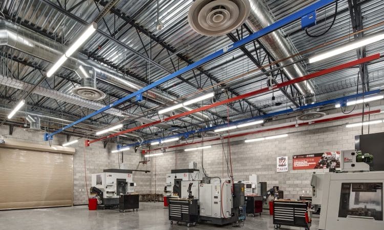 A modern industrial workshop with several CNC machines, workbenches, exposed ductwork, concrete floors, and a large rolling door.