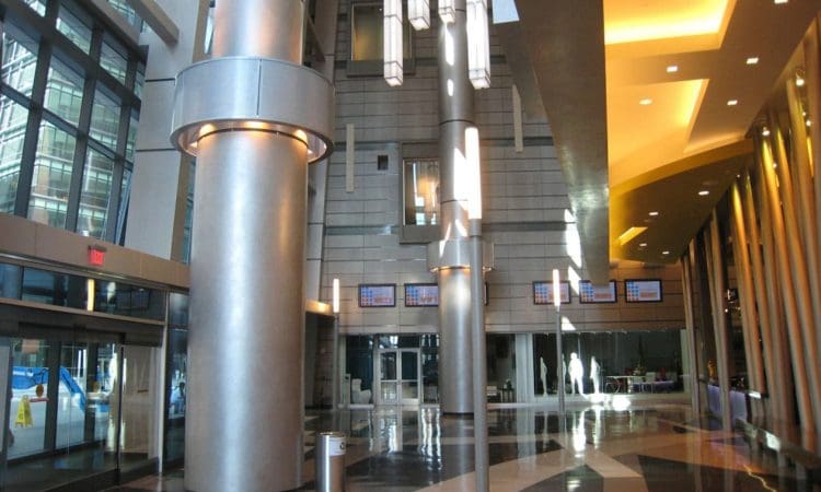 Modern building lobby with large metallic columns, geometric lighting fixtures, shiny patterned floor, and floor-to-ceiling windows letting in natural light.