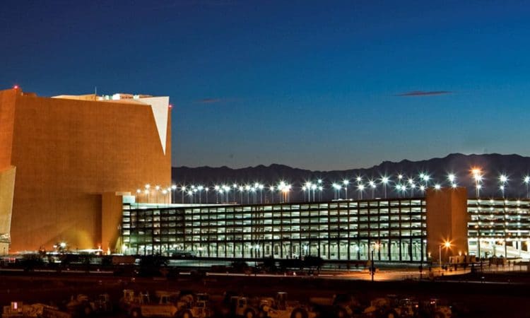 Large, modern building complex with angular architecture and a multi-level parking structure, illuminated at dusk with mountains visible in the background.