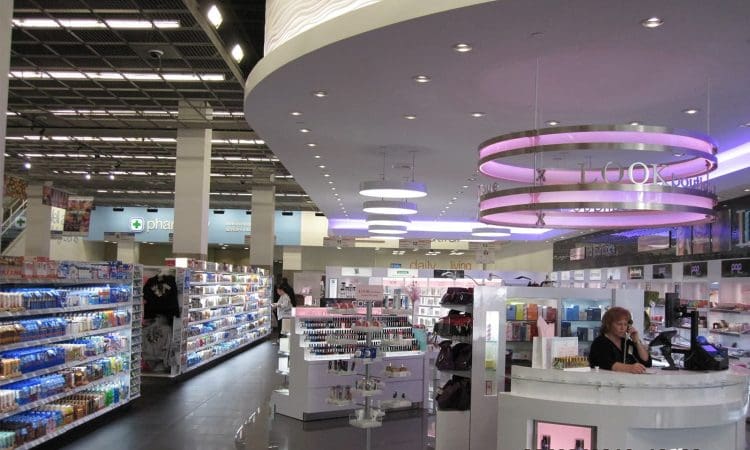 Modern pharmacy interior with shelves of products, a white circular counter with a seated staff member, and overhead lighting. Date and time stamp visible in lower right corner.