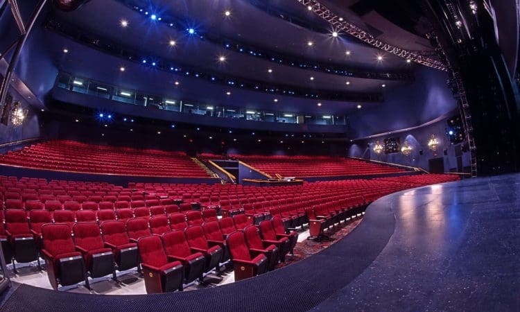 Photo of an empty theater auditorium with multiple tiers of red seats, seen from the stage under blue lighting.