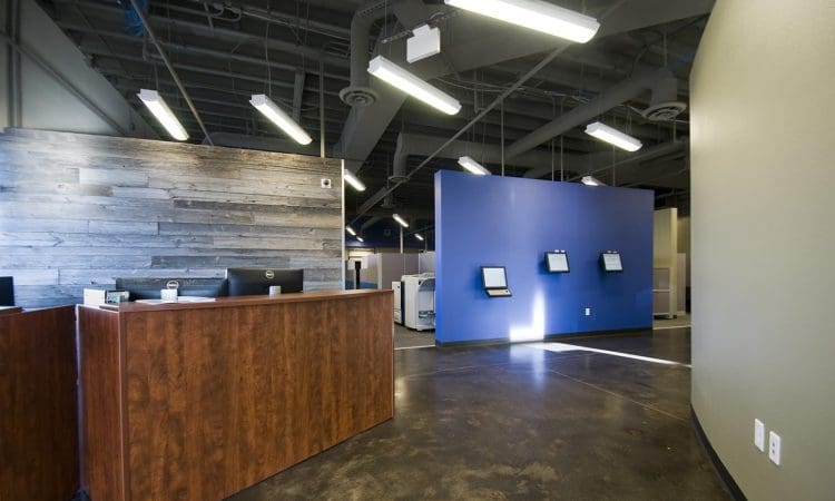 A modern office space with a wooden reception desk, blue accent wall, mounted touchscreens, and exposed ceiling fixtures.