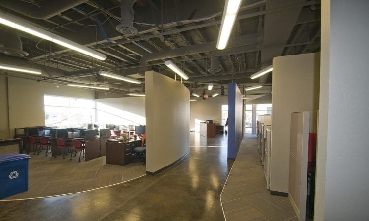 Modern office space with exposed ceiling, curved wall, rows of desks with chairs on the left, cubicles on the right, and bright overhead lighting.
