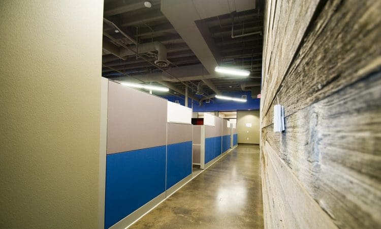 A modern office hallway with polished concrete floors, blue and gray cubicles on the left, and a wooden panel wall on the right under exposed ceiling pipes and lights.