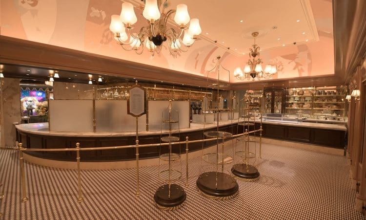 Empty, elegant café interior with chandeliers, marble countertops, and gold stanchions arranged in a queue area; shelves with glassware line the back wall.