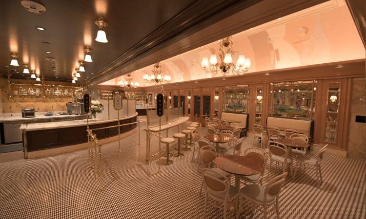 Elegant café interior with marble tables, beige chairs, tiled floor, chandeliers, mirrors, and a counter area; space is empty of people and softly lit.