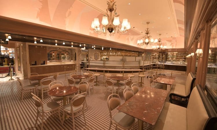 Spacious cafe with round marble tables, beige chairs, chandeliers, and a patterned tile floor under warm lighting. The bar and shelves line the back wall.
