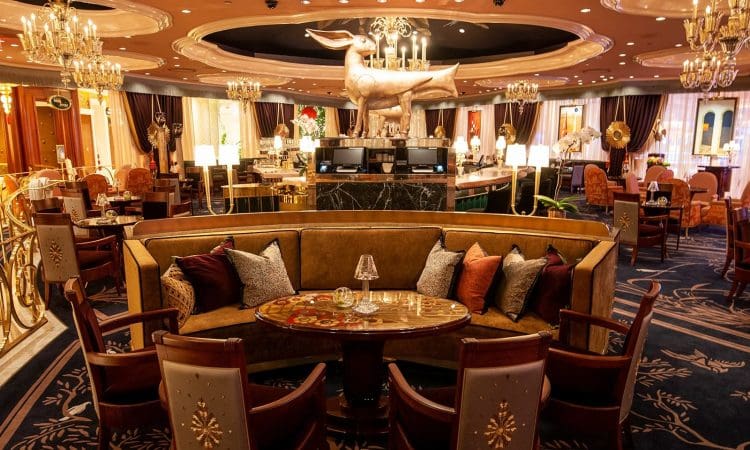 Elegant restaurant interior with plush seating, ornate chandeliers, decorative pillows, a central marble counter, and a large gold dachshund statue above the bar.