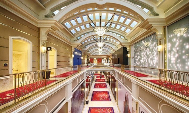 Indoor shopping mall with a high arched glass ceiling, ornate light fixtures, red carpet runners, and luxury storefronts along both sides of the upper and lower walkways.