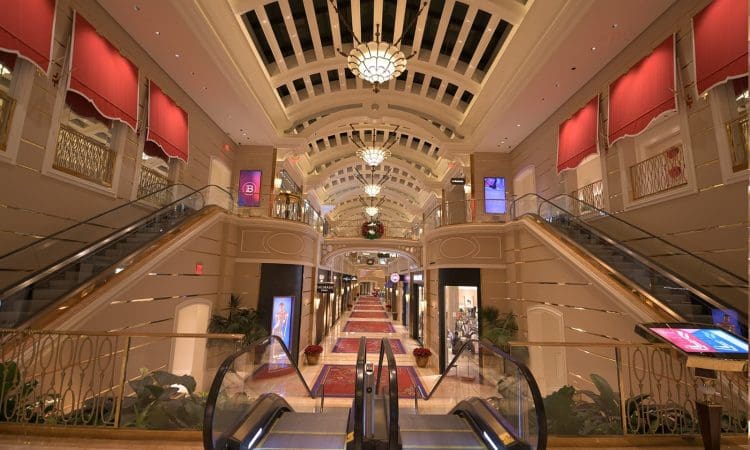 Interior of an upscale shopping mall with chandeliers, red accents, escalators, and various storefronts lining both sides under a vaulted ceiling.