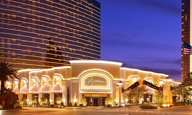 The illuminated entrance of the Wynn hotel and casino in Las Vegas at dusk, with a modern high-rise building and palm trees in the background.