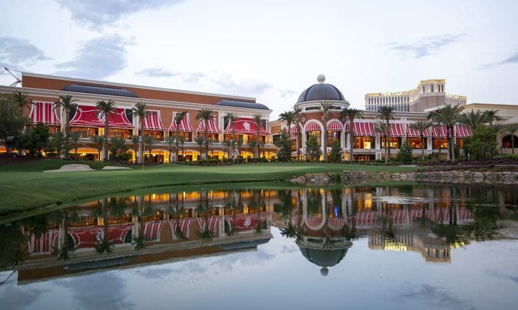 A large building with red-striped awnings and domed roofs is reflected in a calm pond, surrounded by palm trees and grass under a partly cloudy sky.