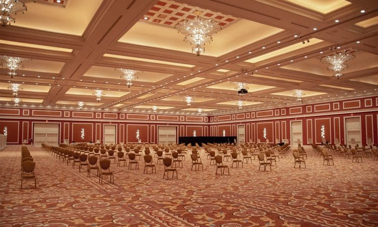 Large, ornate banquet hall with red patterned carpet, chandeliers, and rows of spaced-out chairs facing a small stage at the far end of the room.