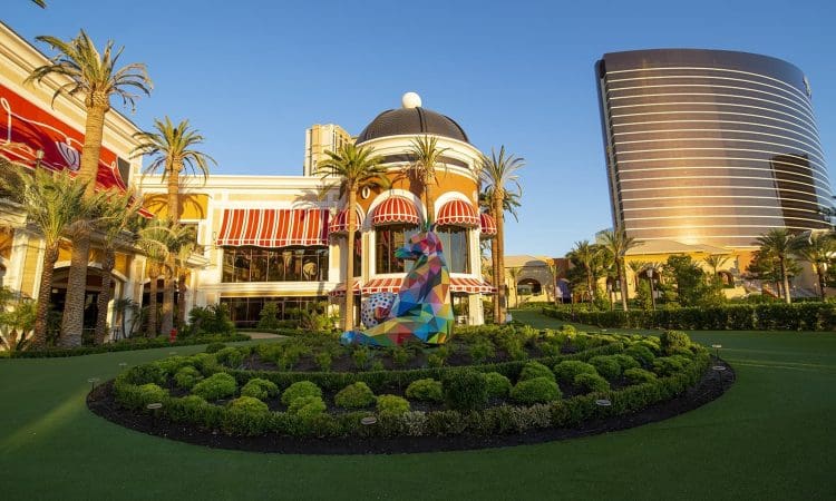Colorful geometric sculpture of a sitting dog on a circular lawn, surrounded by palm trees and modern hotel buildings under a clear blue sky.
