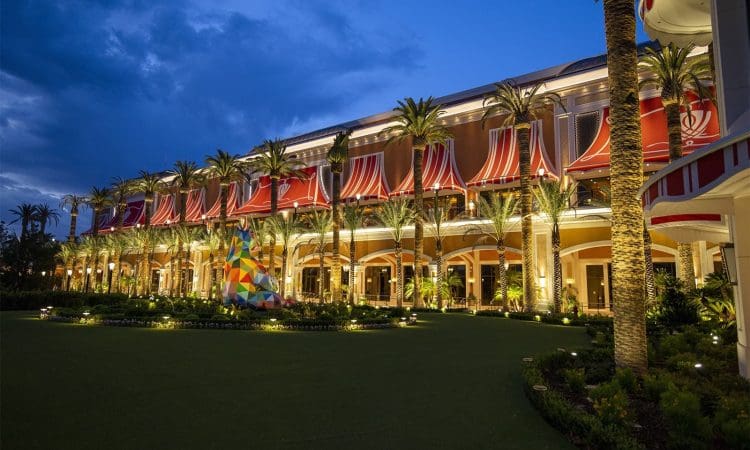 Exterior view of a brightly lit building with red and white awnings, palm trees, and a geometric sculpture on a landscaped lawn at dusk.