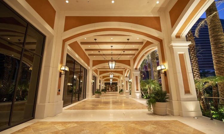 Spacious covered walkway with arched ceilings, large hanging lanterns, tile flooring, and potted plants, leading to a building entrance at night.