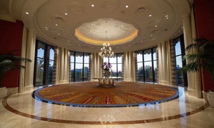 A spacious, elegant lobby with marble floors, a large circular rug, central flower arrangement, chandelier, and tall windows overlooking a scenic view.