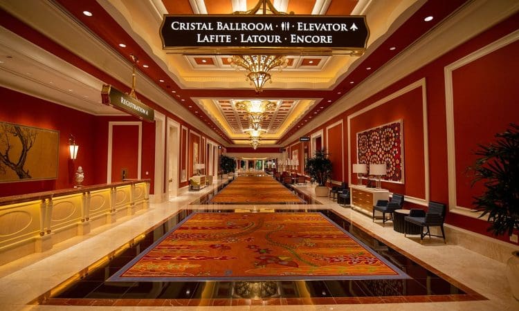 Hotel hallway with red walls, chandeliers, patterned carpet, seating areas, and a ceiling sign directing to ballrooms, elevators, and reception desk.