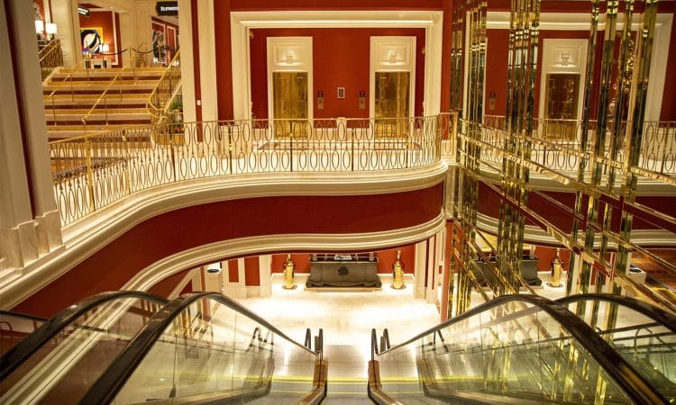 An ornate interior with red walls, gold railings, mirrored panels, and escalators leading down to a lobby area with elevators and stairs.