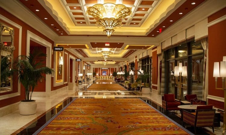 A brightly lit hotel hallway features ornate chandeliers, patterned carpet, seating areas with chairs and tables, potted plants, and large windows on one side.