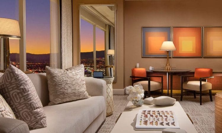 A modern hotel suite with a beige sofa, patterned pillows, a coffee table, a lamp, a dining set for two, and large windows showing a cityscape at sunset.