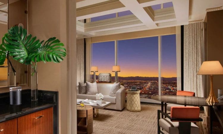 Modern hotel suite with large windows showing a cityscape at sunset, featuring a sofa, desk, lamps, and decorative plants.