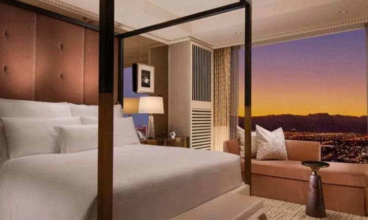 A hotel room with a four-poster bed, a sofa, a side table, and a large window showing a cityscape at sunset.