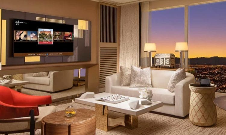 A modern hotel living room features a large TV, neutral-toned furniture, a coffee table, and a large window with a view of a cityscape at sunset.
